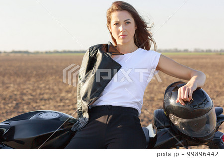 Photo of serious good looking professional female biker dressed in white t shirt and black trousers, has leather jacket on shoulder, keeps hand at helmet, leans on fast motorbike, poses outdoor Photo of serious good looking professional female biker dressed in white t shirt and black trousers, has leather jacket on shoulder, keeps hand at helmet, leans on fast motorbike, poses outdoor 99896442