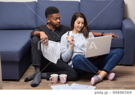 Interracial couple of young people sitting together in front laptop on floor near sofa, in their appartments, busy with composing financial report, using paper documents and portable computer, Interracial couple of young people sitting together in front laptop on floor near sofa, in their appartments, busy with composing financial report, using paper documents and portable computer, 99896569