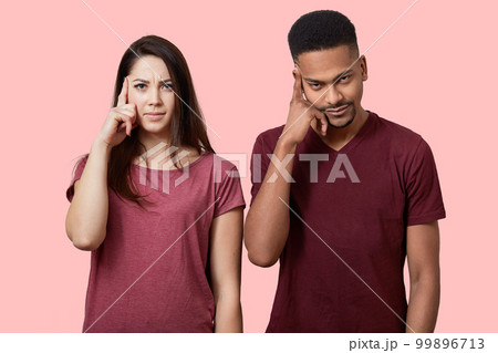 Serious mixed race woman and man keep fore fingers on temple, try to find solution in mind, wear casual clothes, stand nect to each other, isolated over pink background. Relationship and decision Serious mixed race woman and man keep fore fingers on temple, try to find solution in mind, wear casual clothes, stand nect to each other, isolated over pink background. Relationship and decision 99896713