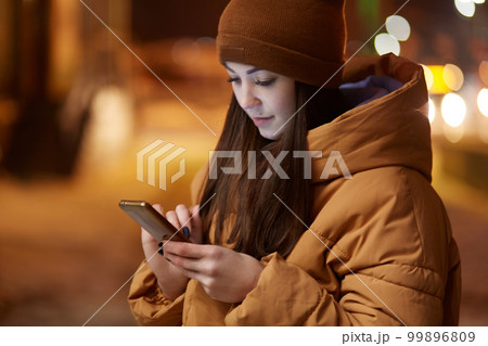 Cropped image of pleasant looking woman concentrated in smart phone, dressed in stylish headgear and jacket, has outdoor walk in evening, searches information in internet. Blurred city view. Cropped image of pleasant looking woman concentrated in smart phone, dressed in stylish headgear and jacket, has outdoor walk in evening, searches information in internet. Blurred city view. 99896809