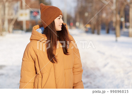 Outdoor shot of brunette young woman focused aside, enjoys winter moments and stroll in open air, dressed in brown stylish headgear and jacket, white snow in background, walks across street. 99896815