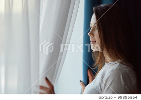 Horizontal shot of attractive brunette pensive young woman in casual white t shirt, looks out of window with thoughtful expression, has make up, enjoys calm domestic atmosphere, poses at her flat. 99896864