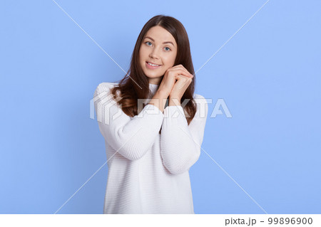 Young cheerful happy girl looking at camera and keeping fists near cheek, posing isolated over blue background, lady with pleasant appearance, woman in white jumper expressing positive emotions. Young cheerful happy girl looking at camera and keeping fists near cheek, posing isolated over blue background, lady with pleasant appearance, woman in white jumper expressing positive emotions. 99896900