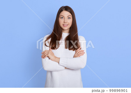 Beautiful young girl posing with folded arms and looking directly at camera, looks happy, expressing confidence, standing isolated over blue background, beautiful lady indoor. Beautiful young girl posing with folded arms and looking directly at camera, looks happy, expressing confidence, standing isolated over blue background, beautiful lady indoor. 99896901