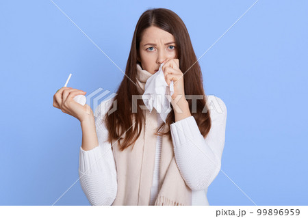 Dark haired unhealthy Caucasian woman with napkin sneezing and holding throat spray in hands to help herself. Rhinitis, cold, sickness and allergy, female uses handkerchief, wearing casual attire. Dark haired unhealthy Caucasian woman with napkin sneezing and holding throat spray in hands to help herself. Rhinitis, cold, sickness and allergy, female uses handkerchief, wearing casual attire. 99896959