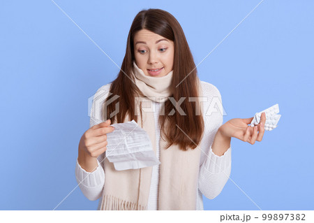 Uncertain pensive young woman holding pills, feeling indecisive about how to treated with them, reading instruction, squeeze shoulders, wearing white casual sweater and scarf. Uncertain pensive young woman holding pills, feeling indecisive about how to treated with them, reading instruction, squeeze shoulders, wearing white casual sweater and scarf. 99897382