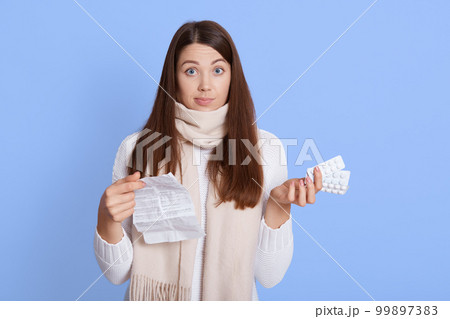 Astonished woman holding pills with instruction and looking at camera with puzzled facial expression, don't know how to take medicine, wearing white sweater and scarf, isolated over blue background. 99897383