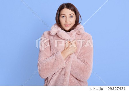 Frozen young brown hair woman posing isolated over blue background, portrait, wrapping herself in pale pink fur coat, needs warm up, looks directly at camera. Frozen young brown hair woman posing isolated over blue background, portrait, wrapping herself in pale pink fur coat, needs warm up, looks directly at camera. 99897421
