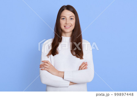 Happy smiling young woman dresses white shirt, having dark hair, looking directly at camera, keeps hands folded, expressing positive, standing isolated over blue background. Happy smiling young woman dresses white shirt, having dark hair, looking directly at camera, keeps hands folded, expressing positive, standing isolated over blue background. 99897448