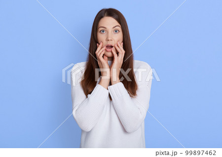 Stunned caucasian female stares at camera with big eyes and keeps mouth opened, looks surprisingly, realizes bad news, stands against blue background, keeps fingers on cheeks. 99897462