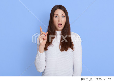 Amazed shocked young woman pointing up with fore finger, keeps mouth widely opened, wearing casual white shirt, has dark hair, being shocked and astonished, stands against blue wall. 99897466
