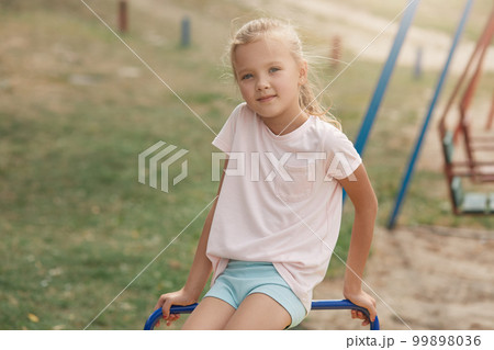Photo of little girl on playground, spending time with friends and playing outdoors in summer. Happy kid in kindergarten or preschool having fun, looking at camera, dressed casualt shirt and short. 99898036