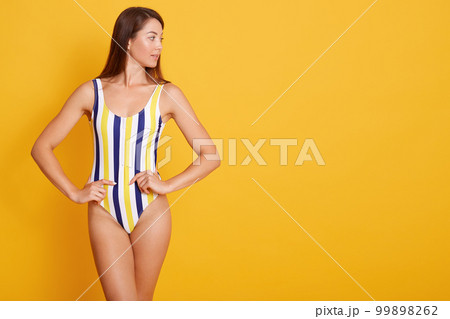 Photo of young woman with straight dark hair dressed in swimsuit, looking aside isolated over yellow background in studio, keeps hjands on hips, copy space for advertismant or promotion text. Photo of young woman with straight dark hair dressed in swimsuit, looking aside isolated over yellow background in studio, keeps hjands on hips, copy space for advertismant or promotion text. 99898262