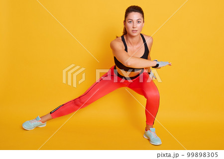 Studio shot of young beautiful woman working out, doing side lunges, slimledy exercises for legs, hips and buttocks, sporty female wears black top and leggins looks at camera, isolated on yellow wall. 99898305
