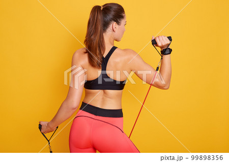 Picture of attarctive sporty woman performs exercising with resistance band, wearing black topand leggins,model posingisolated over yellow background. Sport, strength and motivation concept.. Picture of attarctive sporty woman performs exercising with resistance band, wearing black topand leggins,model posingisolated over yellow background. Sport, strength and motivation concept.. 99898356