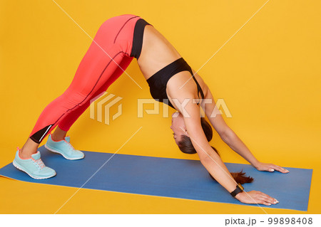 Woman do yoga Downward Facing Dog pose insolated over yellow studio background, beautiful slim and sporty female strengthening back exercise on yoga mat at home. Healthy lifestyle and sport concept. Woman do yoga Downward Facing Dog pose insolated over yellow studio background, beautiful slim and sporty female strengthening back exercise on yoga mat at home. Healthy lifestyle and sport concept. 99898408