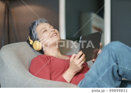 Mature woman with grey hair is depicted listening to music while sitting on sofa in her home. She has her eyes closed and is holding a tablet in her hands. High quality photo Mature woman with grey hair is depicted listening to music while sitting on sofa in her home. She has her eyes closed and is holding a tablet in her hands. High quality photo 99899831