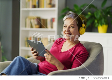 Mature woman smiles on a sofa, holding tablet PC and listening to music on headphones. She enjoys leisure, technology and music, feeling tranquil and blissful. High quality photo Mature woman smiles on a sofa, holding tablet PC and listening to music on headphones. She enjoys leisure, technology and music, feeling tranquil and blissful. High quality photo 99899864