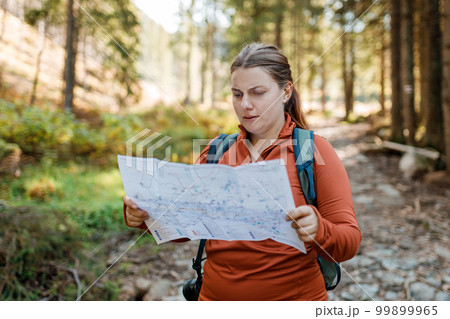 Sad female tourist is exploring new locations on forest background. Redhead girl holding a paper map in a sunny day. Travel concept. Hiking woman wearing knit hat exercising outdoor Sad female tourist is exploring new locations on forest background. Redhead girl holding a paper map in a sunny day. Travel concept. Hiking woman wearing knit hat exercising outdoor 99899965