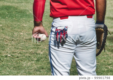 A baseball player at a match wearing a leather mitt and holding a ball in his hand, with a small glove sticking out of his back pocket in white pants, standing on the grass A baseball player at a match wearing a leather mitt and holding a ball in his hand, with a small glove sticking out of his back pocket in white pants, standing on the grass 99901798