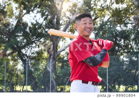 A young Asian man holding a baseball bat standing in a ballpark smiling A young Asian man holding a baseball bat standing in a ballpark smiling 99901804