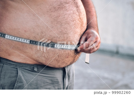 Close-up of a man's round and full belly, checking the width of his stomach with a measuring tape in centimeters Close-up of a man's round and full belly, checking the width of his stomach with a measuring tape in centimeters 99902260