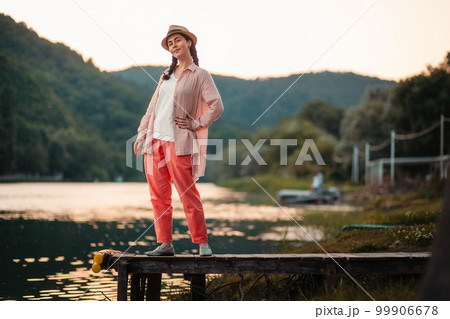 Summer vacation. A stylish pretty woman in summer clothes and straw hat posing stands at river pier. Copy space. Beautiful landscape at the background Summer vacation. A stylish pretty woman in summer clothes and straw hat posing stands at river pier. Copy space. Beautiful landscape at the background 99906678