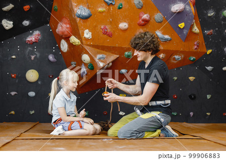 Instructors man are helping client small girl to put on safety harnesses in rock climbing center indoors. Little girl is engaged in rock climbing in the children's entertainment center. Blonde 99906883