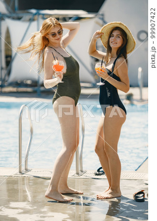 Caucasian girls in swimwear and sunglasses are drinking cocktails, talking and smiling. Female friends standing near swimming pool. Girls wearing black and khaki swimwear. 99907422
