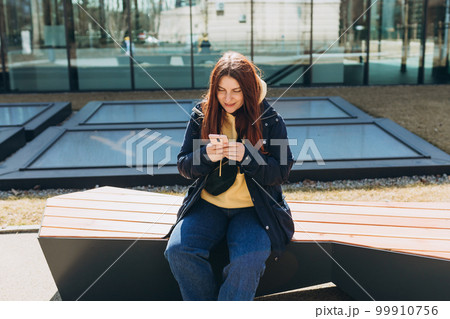 Beautiful woman in casual clothing using her smart phone while sitting on the bench on city street. Happy Young Middle Eastern woman sitting on a bench after college classes. Urban lifestyle concept 99910756
