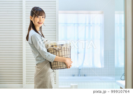 バスルームで洗濯物を運ぶエプロンを着た笑顔の若い主婦 99910764