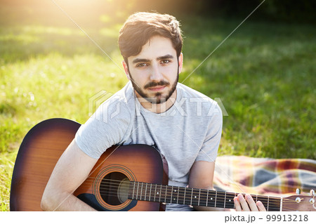 Young bearded male guitarist in casual T-shirt sitting with musical instrument on green grass, playing different songs, looking at camera with serious look, resting at nature. People, hobby, music Young bearded male guitarist in casual T-shirt sitting with musical instrument on green grass, playing different songs, looking at camera with serious look, resting at nature. People, hobby, music 99913218