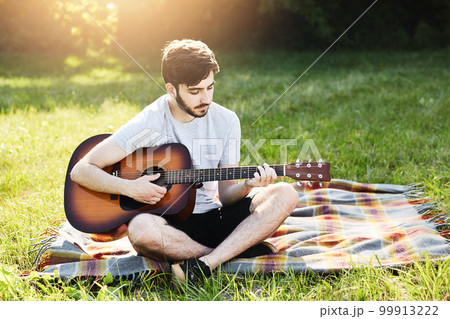 Portrait of attractive stylish bearded guy with guitar sitting crossed legs on green grass, playing musical instrument, enjoying calm atmosphere. Talented boy demonstrating his talent while resting Portrait of attractive stylish bearded guy with guitar sitting crossed legs on green grass, playing musical instrument, enjoying calm atmosphere. Talented boy demonstrating his talent while resting 99913222