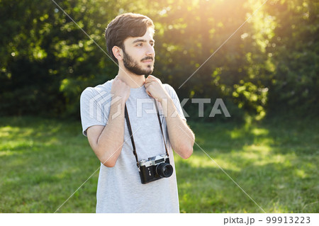 Portrait of stylish bearded man with retro camera standing at green park looking into distance with thoughtful expression. Young male photographer making photos of beautiful landscapes outdoor Portrait of stylish bearded man with retro camera standing at green park looking into distance with thoughtful expression. Young male photographer making photos of beautiful landscapes outdoor 99913223