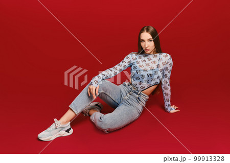 Young girl in transparent top and jeans sits on the floor in red background Young girl in transparent top and jeans sits on the floor in red background 99913328