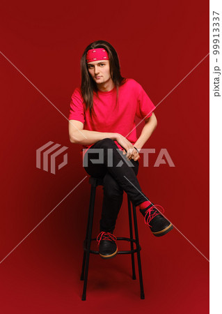 Young man sits on high stool with his leg on another leg and leaning forward 99913337