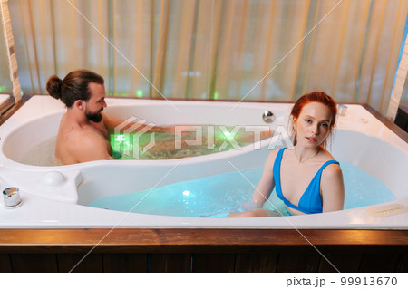 Attractive young woman in bikini and brutal bearded man sitting in double hot bath tub with hydromassage at luxury spa salon, enjoying weekend in bathtub. Romantic relationship, passionate honeymoon. 99913670