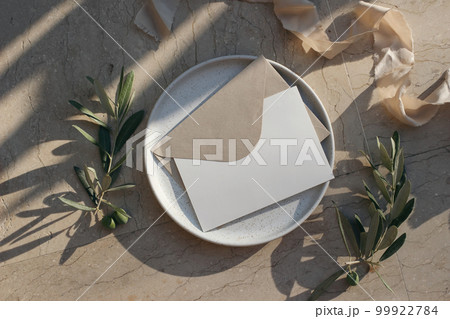 Summer branding, wedding stationery. Blank greeting card, envelope mock up on ceramic plate in sunlight. Beige marble background. Olive tree branches, fruit, ribbon. Long shadows. Flat lay, top view. 99922784