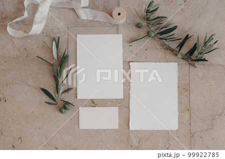 Italian summer wedding stationery set. Mock-up scene with blank paper greeting, RSVP cards on beige marble tiles background. Green olive tree branches, silk ribbon. Mediterranean flat lay, top view. 99922785