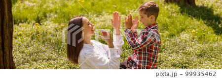 Happy joyful mother play with little son and clapping in hands while sitting on grass in park. 99935462