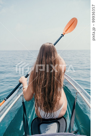 Woman in kayak back view. Happy young woman with long hair floating in transparent kayak on the crystal clear sea. Summer holiday vacation and cheerful female people relaxing having fun on the boat Woman in kayak back view. Happy young woman with long hair floating in transparent kayak on the crystal clear sea. Summer holiday vacation and cheerful female people relaxing having fun on the boat 99936357