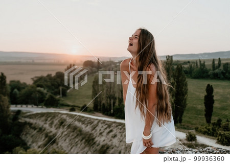 Happy woman standing with her back on the sunset in nature in summer with open hands. Romantic beautiful bride in white boho dress posing with mountains on sunset 99936360