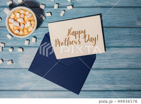 HAPPY MOTHER'S DAY text on card inscription positive quote phrase Greeting card blue envelope with white cup of coffee and marshmallows on wooden blue background holiday HAPPY MOTHER'S DAY text on card inscription positive quote phrase Greeting card blue envelope with white cup of coffee and marshmallows on wooden blue background holiday 99936576
