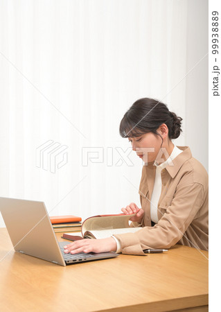 英文の古書を読みつつノートパソコンで調べ物をする若い女性 99938889