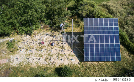 Top view of male workers installing pole mounts for photovoltaic solar panels on sunny day. Three men mounting supporting structures for solar modules in the field with green trees. 99943386