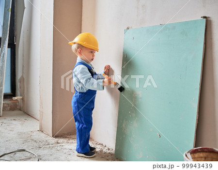 Little boy construction worker painting wall with brush in apartment under renovation. Child wearing safety helmet and work overalls while playing at home. 99943439