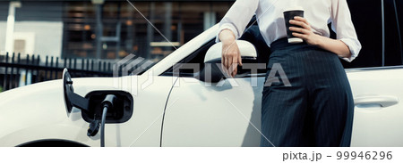 Progressive woman with coffee while charging EV car with residential buildings. 99946296