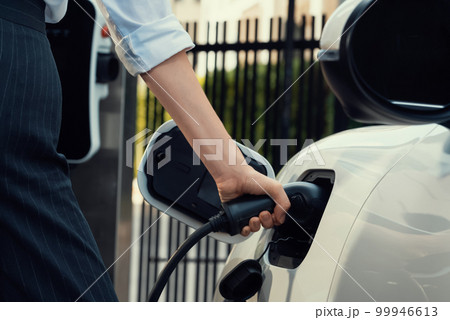 Focus EV charger plug with blur progressive woman charge EV car in background. 99946613