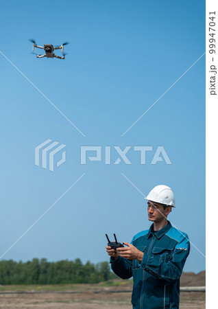 A man in a helmet and overalls controls a drone at a construction site. The builder carries out technical oversight. A man in a helmet and overalls controls a drone at a construction site. The builder carries out technical oversight. 99947041