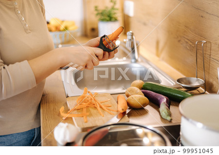 Female peel carrot using vegetable peeler. Close-up of woman's hand at kitchen. Preparing food at home 99951045